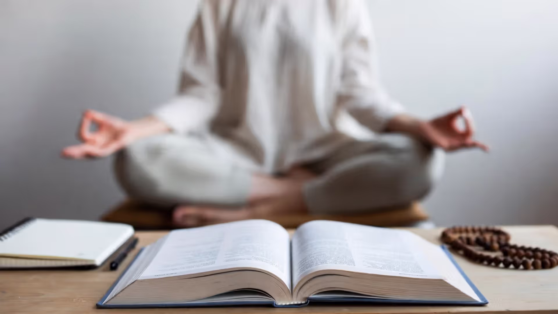 Open book labeled “Yoga Sutras” on a desk with a person meditating in the background.